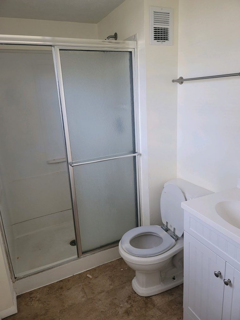 a standard plain white bathroom with a shower, sink, and toilet occupying the same small room